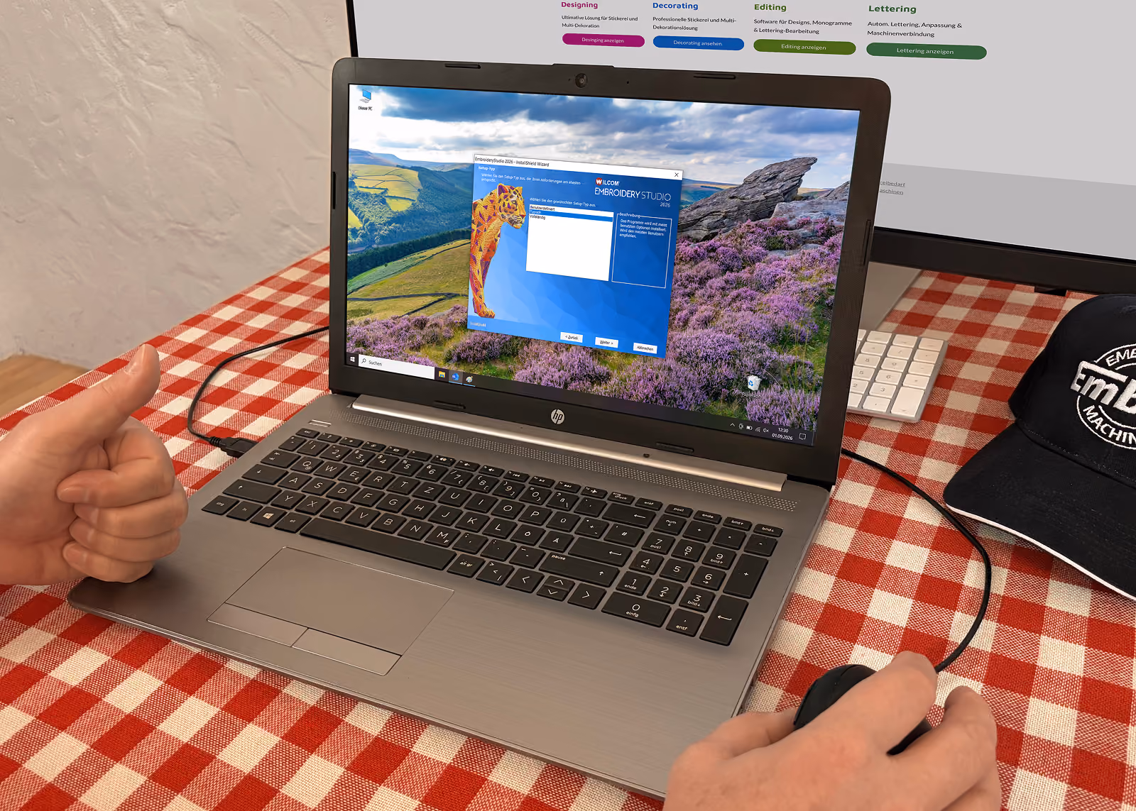 Laptop auf einem rot-weiß karierten Tischtuch mit einer Installationssoftware für V7.60 WILCOM EMBROIDERY STUDIO auf dem Bildschirm; eine rechte Hand benutzt eine Maus, eine linke zeigt Daumen hoch.