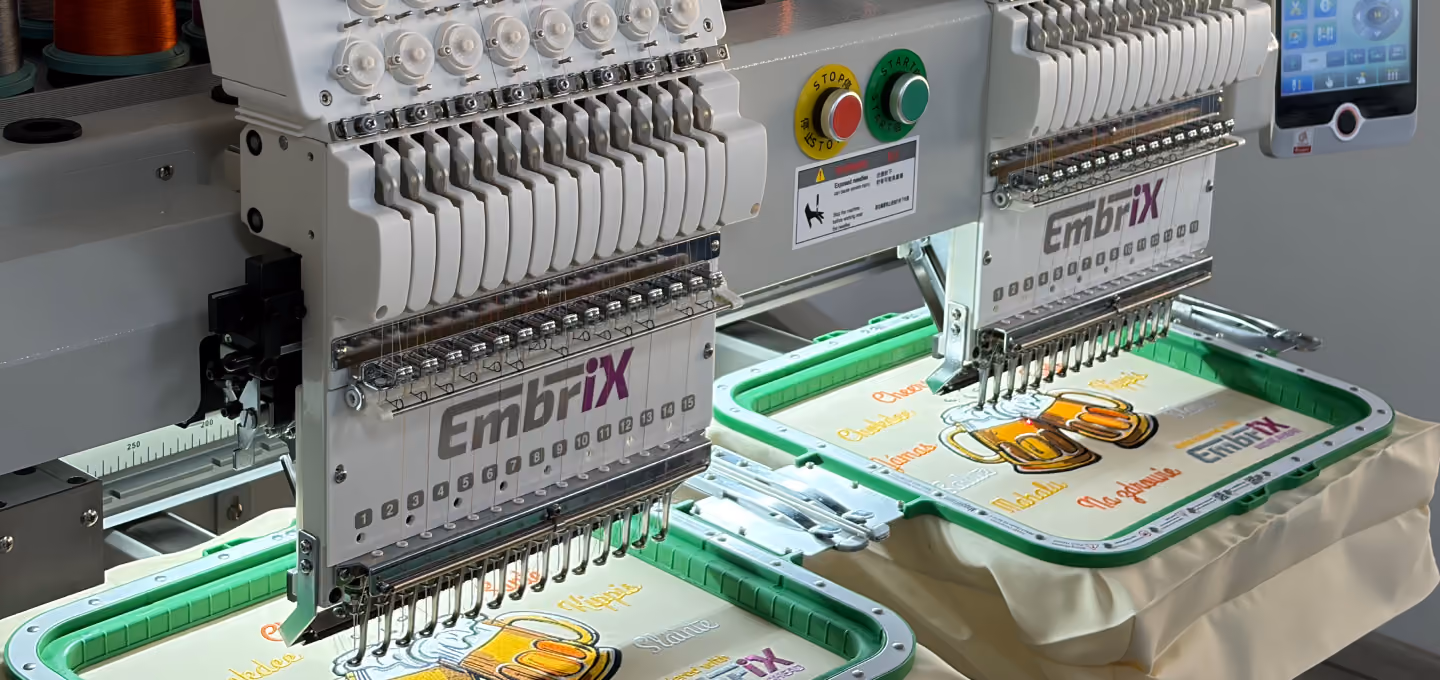 Zweistufige industrielle Stickmaschine mit der Marke EmbriX, die hell beleuchtete gelbe und orangefarbene Designs auf weißen Stoffen stickt.