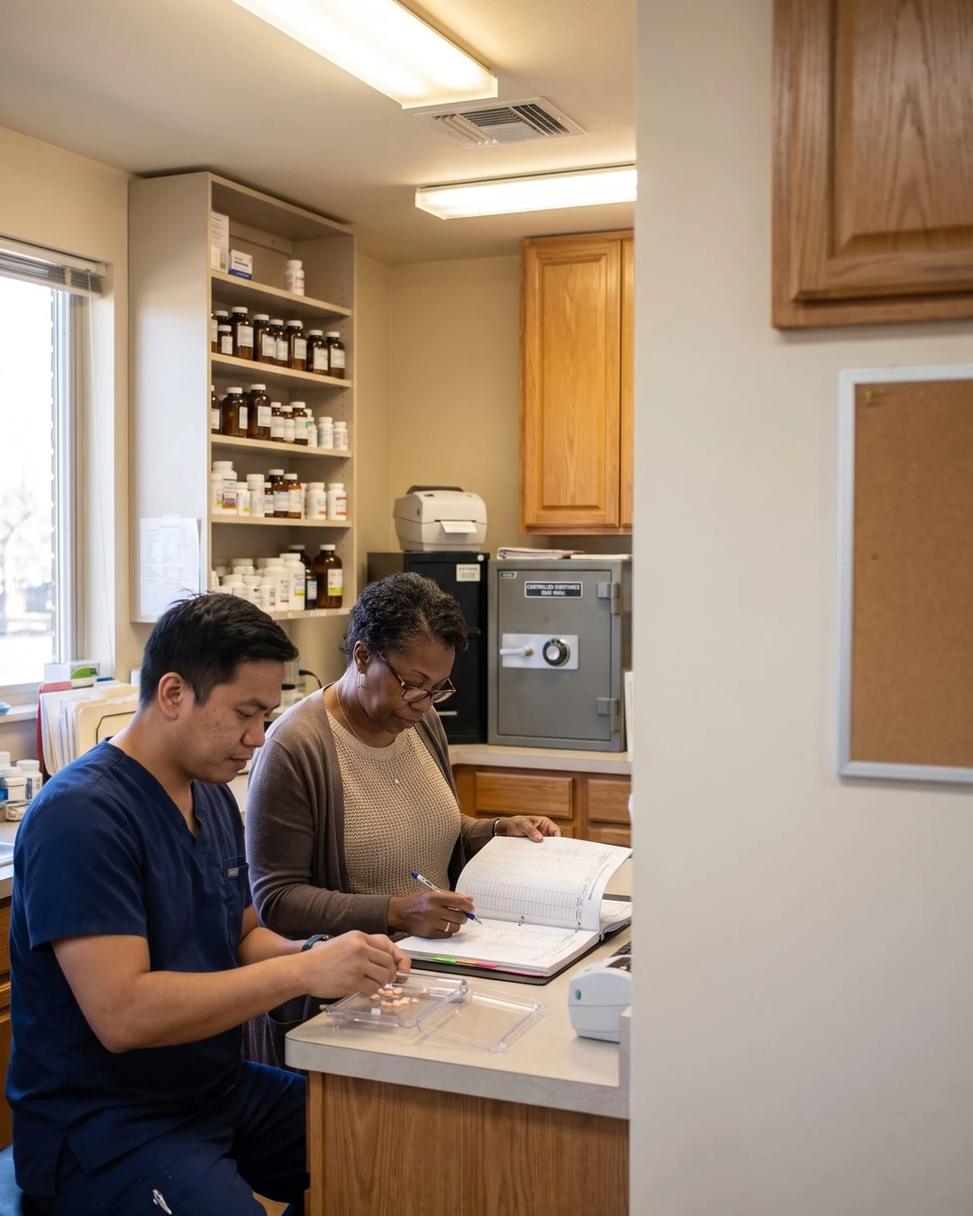A technician and a manager review a controlled-substance log beside the clinic pharmacy safe.