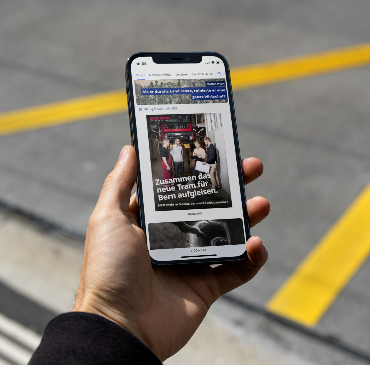 Hand hält ein Smartphone mit einer Anzeige der BERNMOBIL Kampagne, im Hintergrund Strassenmarkierungen