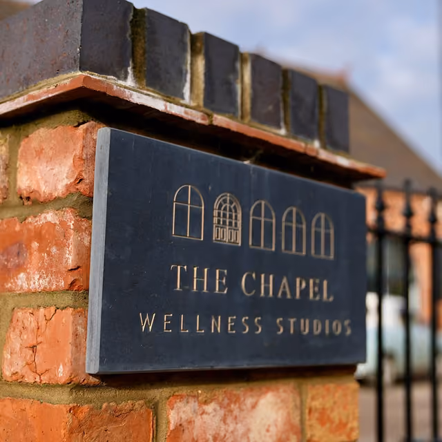 Black rectangular sign mounted on a brick pillar with gold lettering and window icons, reading 'THE CHAPEL WELLNESS STUDIOS'.