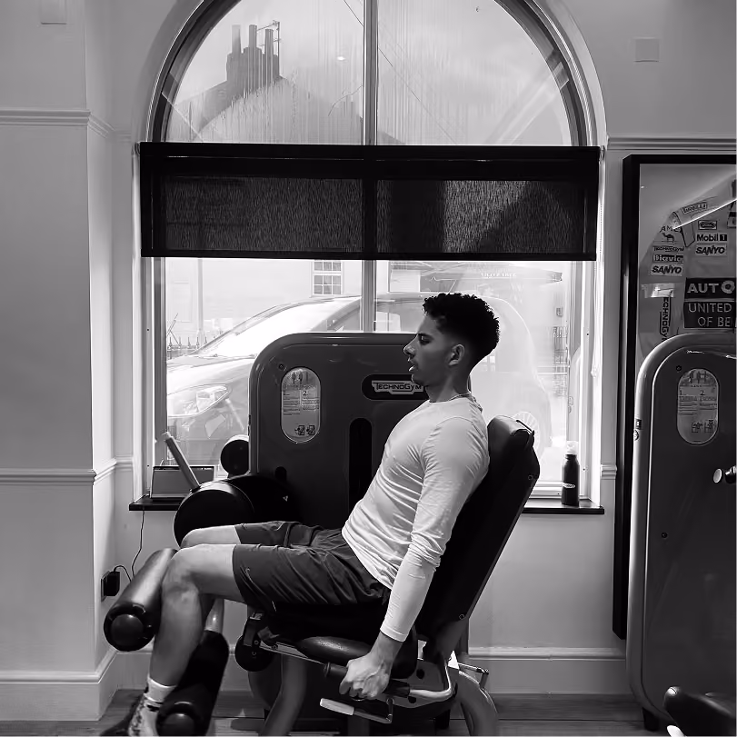 A man in a long-sleeve shirt and shorts seated on a leg extension gym machine exercising his legs in front of a large arched window with partially lowered blinds.