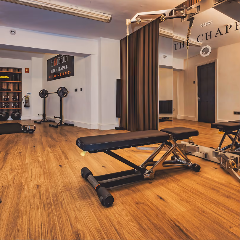 Interior view of a wellness studio with wooden flooring, a black adjustable workout bench in the center, mirrors on the right wall, and various fitness equipment including kettlebells and medicine balls on shelves along the left wall, with a sign reading 'THE CHAPEL WELLNESS STUDIOS' on the back wall.