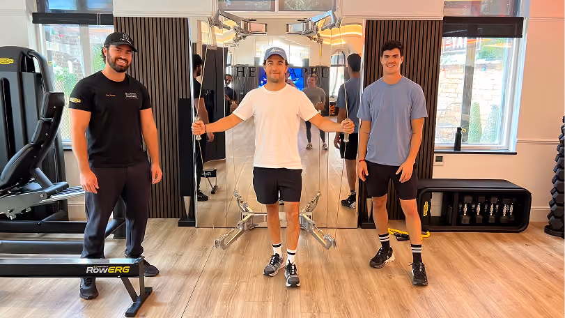 Three young men standing in a gym with wooden floors and large windows; the center man holds cable attachments from a fitness machine, and the others stand on either side, smiling.