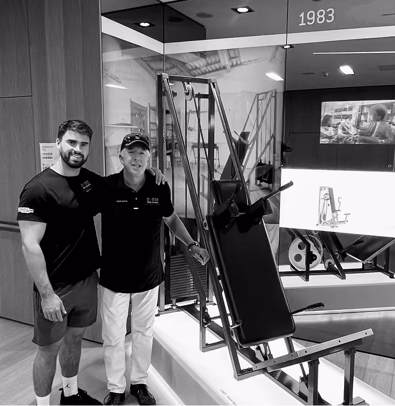 Two men standing side by side with one arm around the other next to a gym machine displayed in a modern showroom with screens and the year 1983 above.