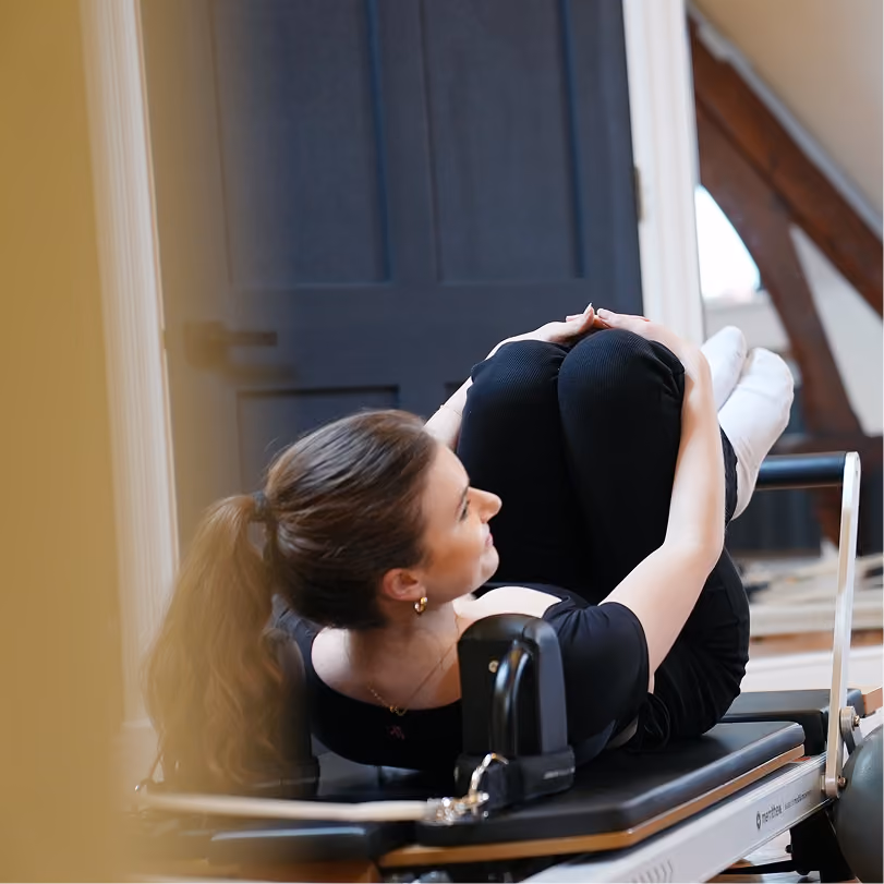 Young woman with a ponytail practicing Pilates on a reformer machine, holding her knees close to her chest indoors near a dark door and wooden beams.