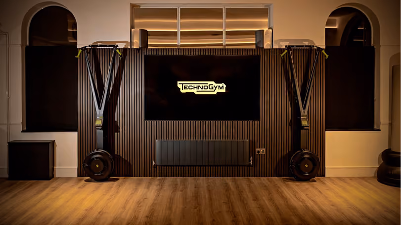 Indoor gym space with wooden floor, two pieces of fitness equipment on each side, and a large screen in the center displaying the TechnoGym logo, with two arched windows on the wall behind.