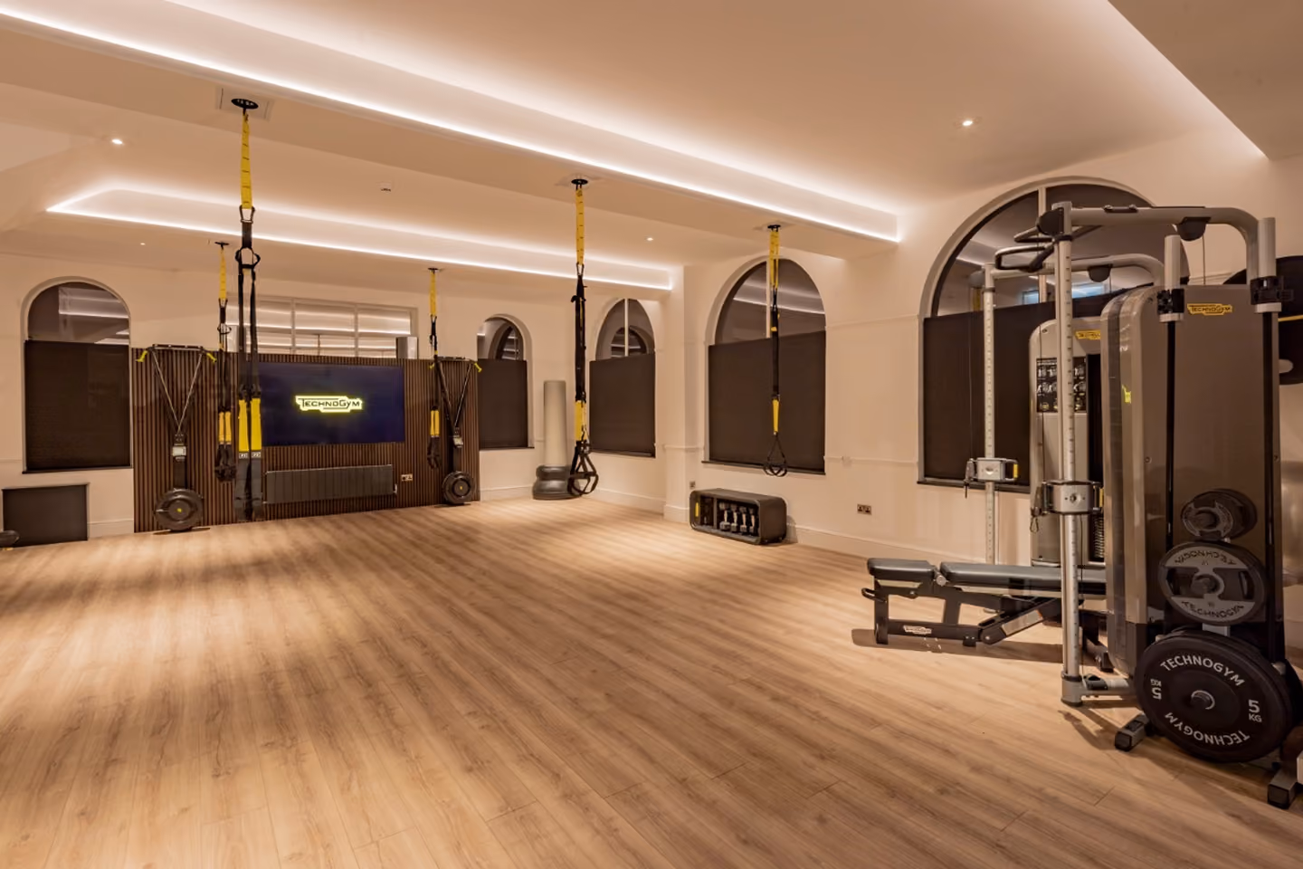 Spacious modern gym studio with wooden floor, arched windows with black shades, ceiling LED strip lighting, yellow and black suspension training straps, and various exercise machines and weights.