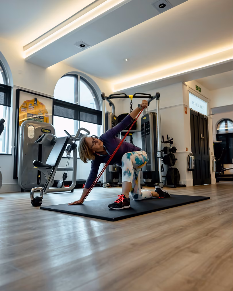 Woman exercising on a black mat in a gym, stretching with a red resistance band while kneeling on one leg, surrounded by gym equipment and large windows.