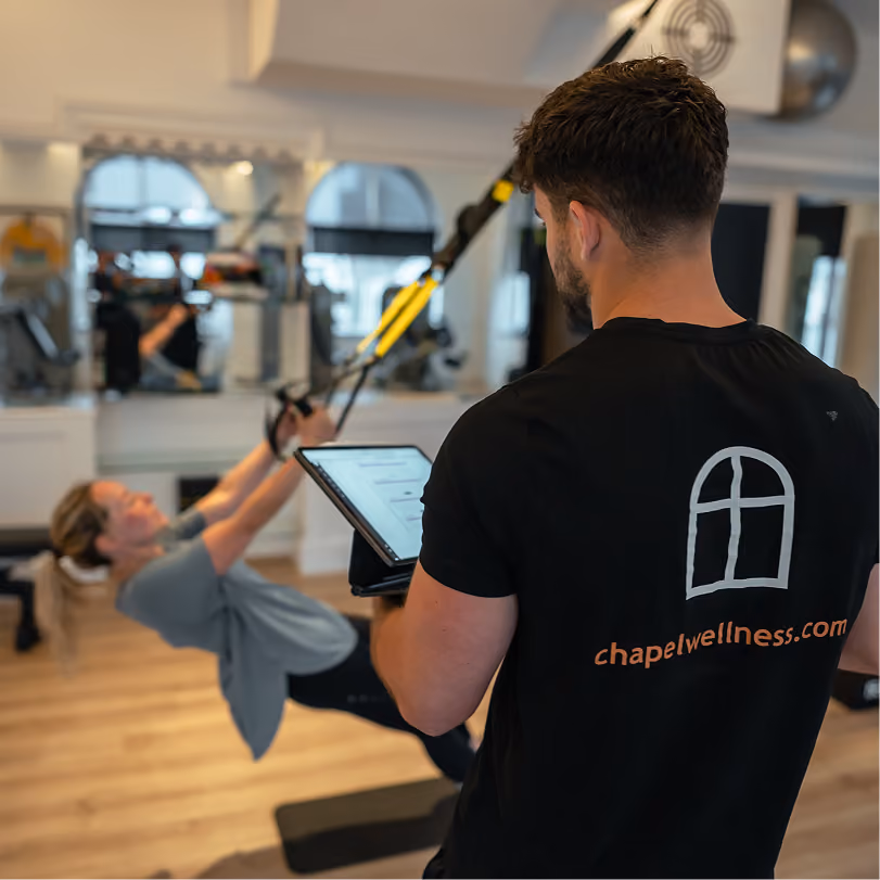 A male trainer wearing a black chapelwellness.com shirt monitors a woman performing suspension training exercises with resistance bands in a gym with wooden floors and mirrors.