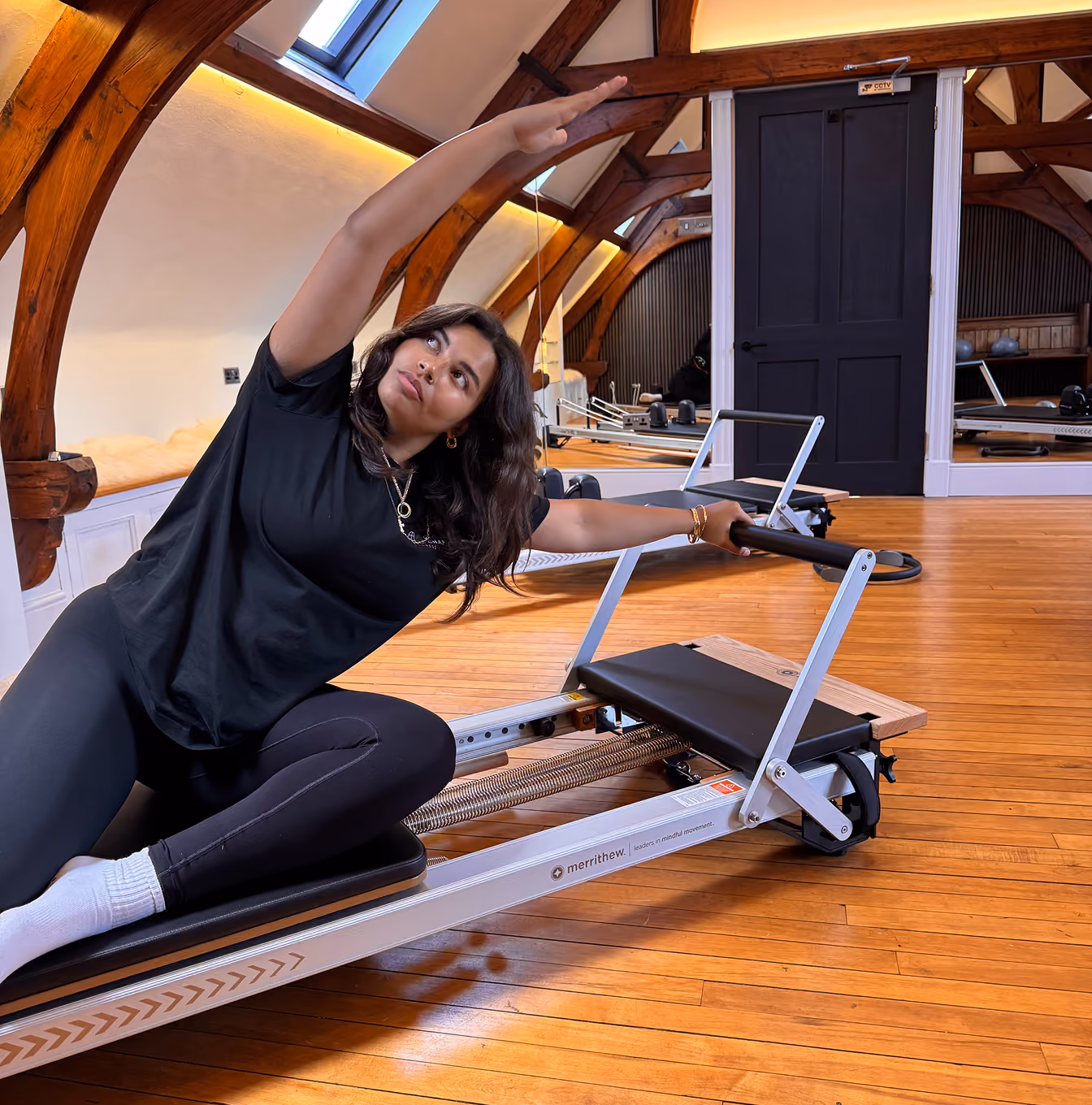 Woman performing a side stretch on a Pilates reformer machine in a spacious room with wooden floors and arched wooden beams.