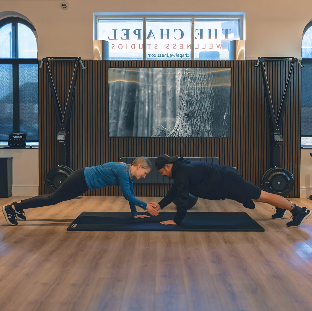 A woman and a man facing each other in a plank position on black mats in a fitness studio, clasping one hand for a partner workout.