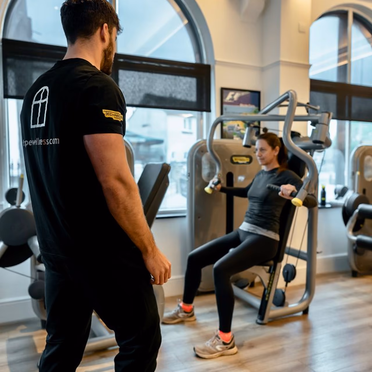 A gym trainer watches a woman using a chest press machine in a bright gym with large windows and wooden floors.