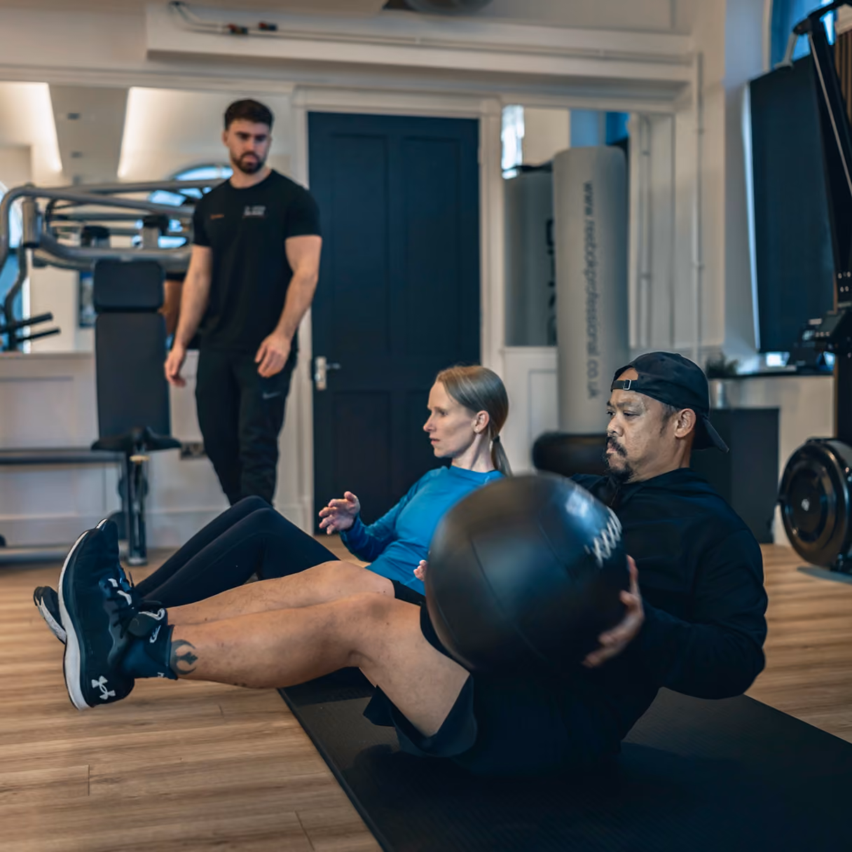 Two people exercising on mats in a gym, performing seated abdominal exercises with a medicine ball while a trainer in black watches them.