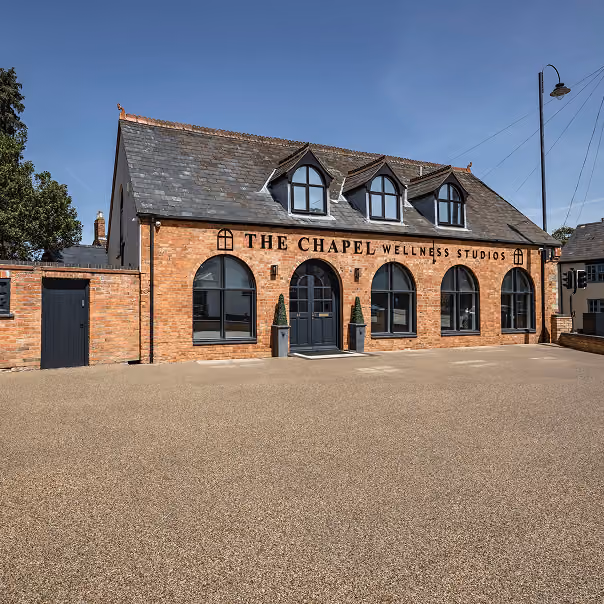 Brick building with a slate roof and three dormer windows labeled 'THE CHAPEL WELLNESS STUDIOS' above four large arched windows and a central door, with two potted plants by the entrance under a clear blue sky.