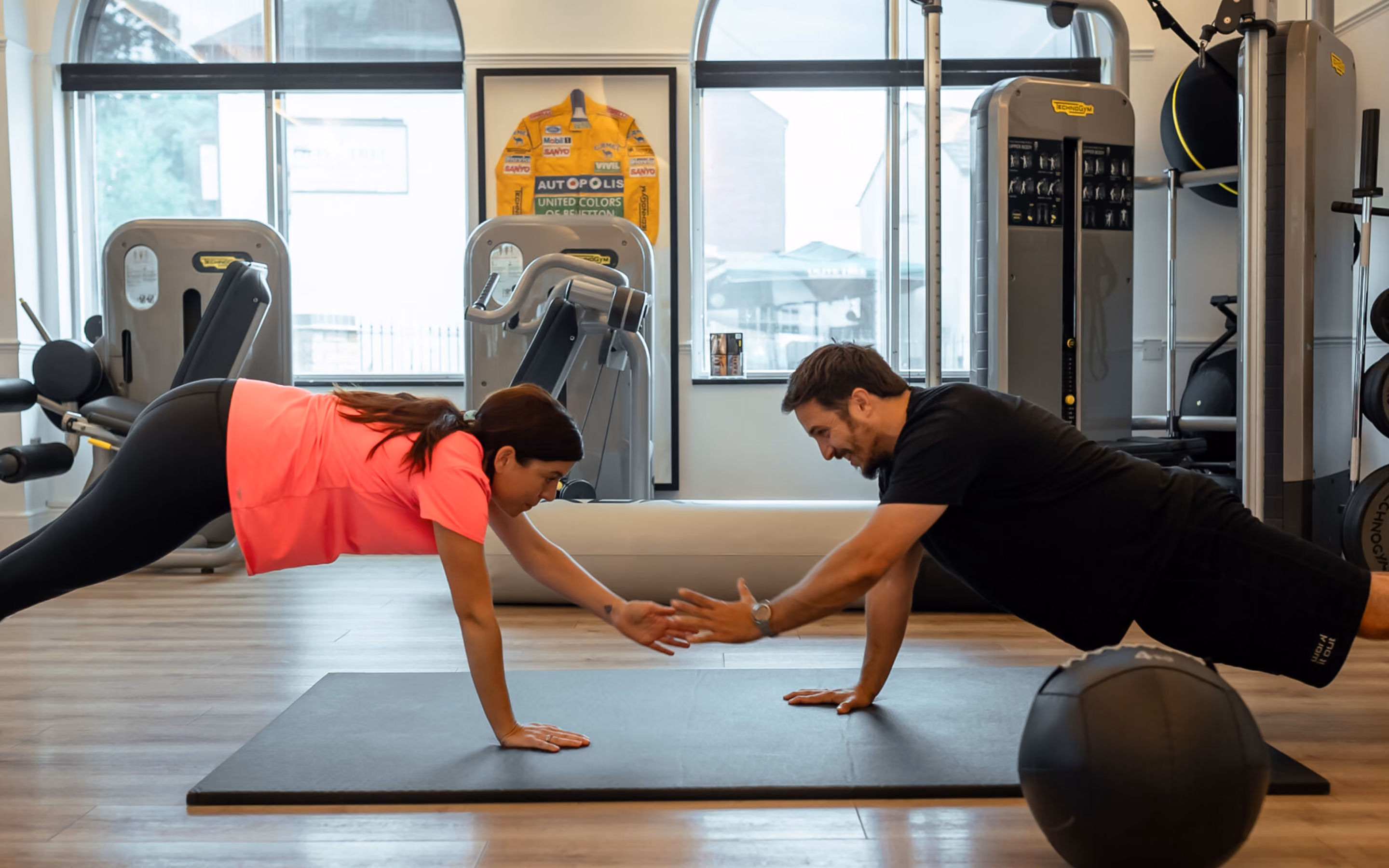 A man and woman in a gym performing a plank exercise facing each other while reaching out to touch hands over a black mat, surrounded by workout equipment and large windows.