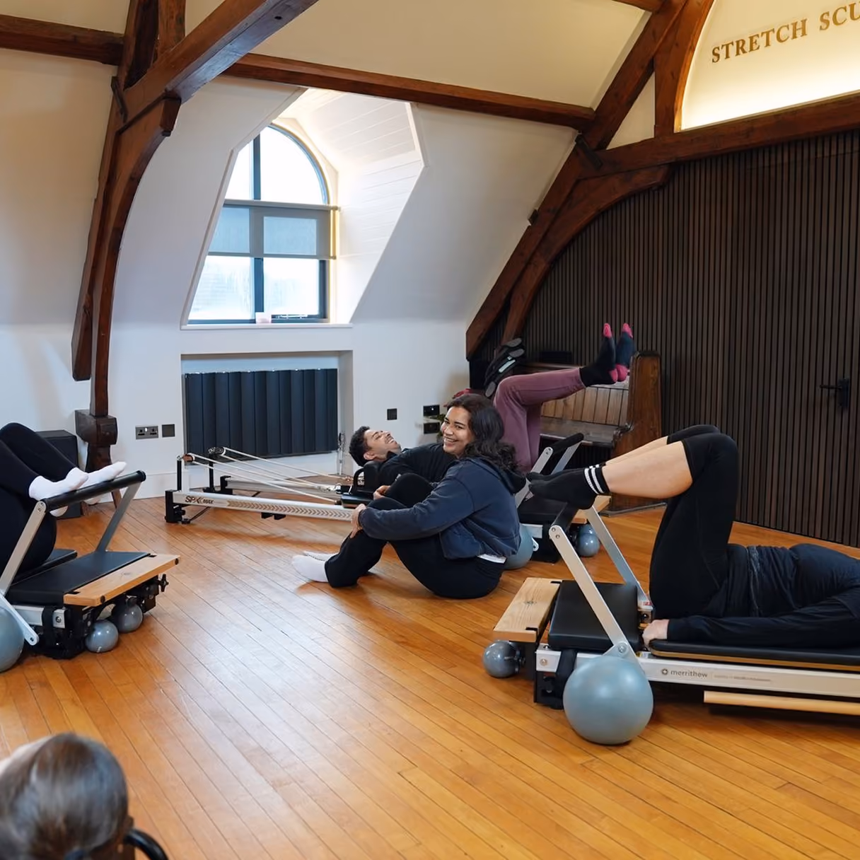 Group of people exercising on Pilates reformer machines in a bright room with wooden beams and a large arched window, stretching and smiling.
