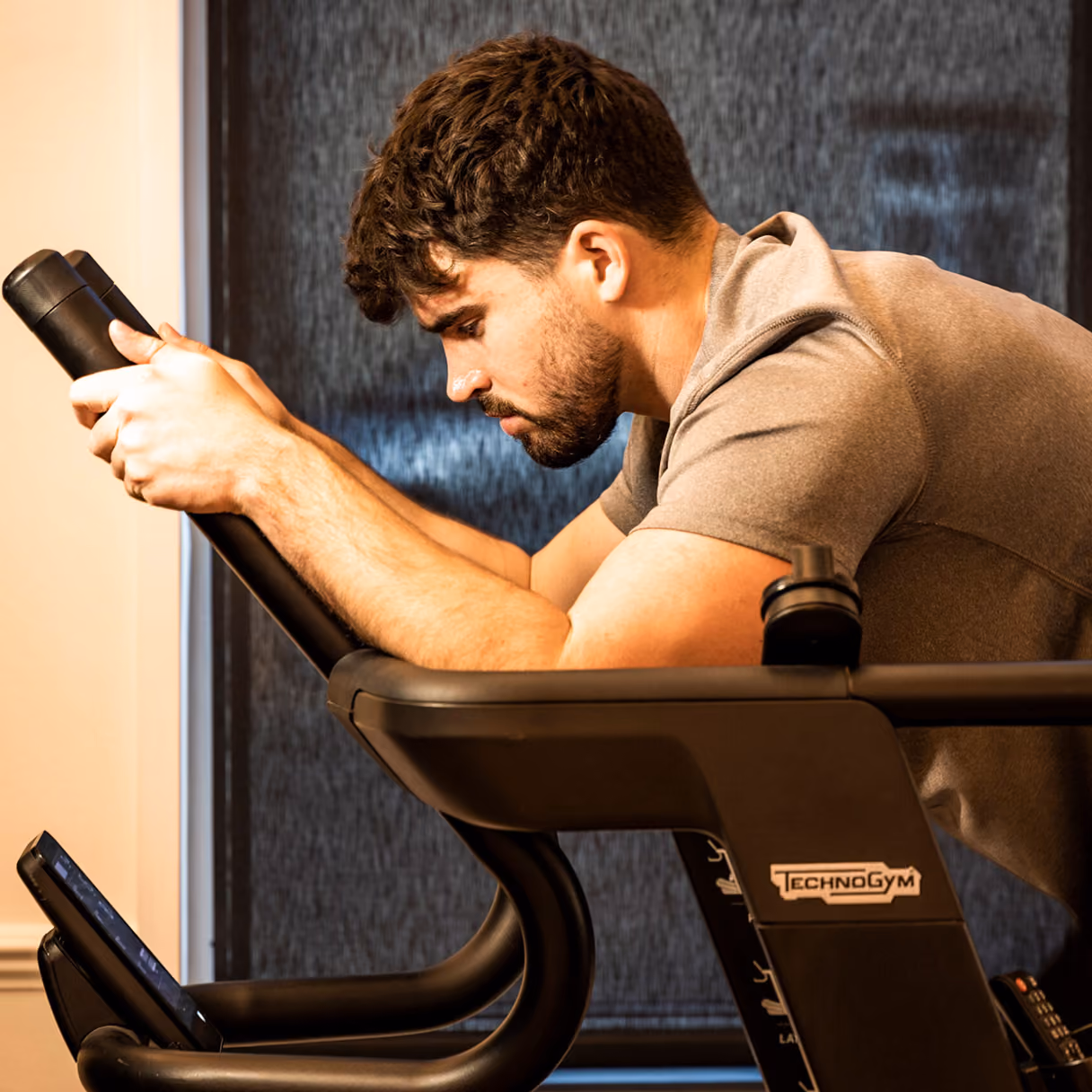 Young man with short curly hair and beard leaning forward holding handles on a Technogym exercise bike, looking focused at the digital screen.