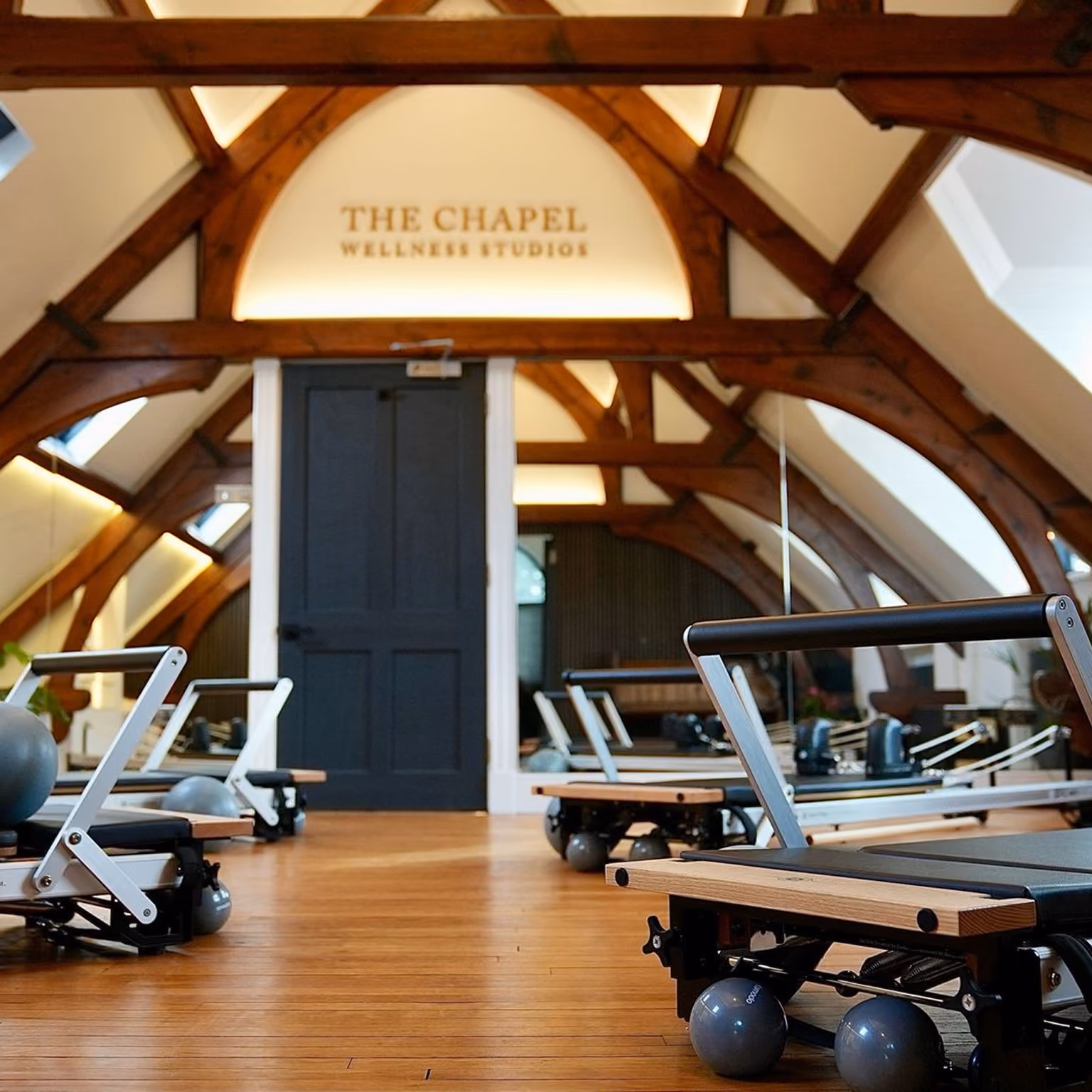 Interior of The Chapel Wellness Studio with wooden floor, exposed wooden beams, a black door, mirrored walls, and Pilates reformer machines arranged in the room.