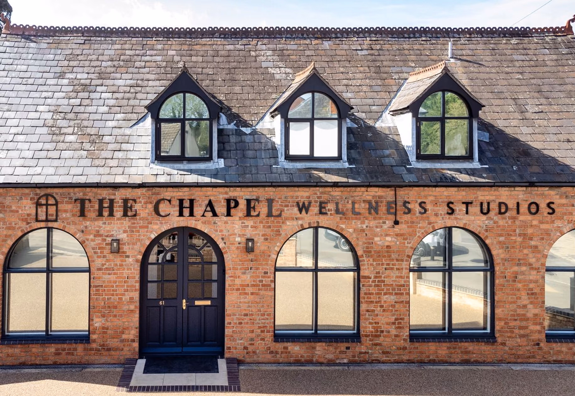 Exterior view of The Chapel Wellness Studios building with a brick facade, black framed arched windows and door, and a slate roof with three dormer windows.
