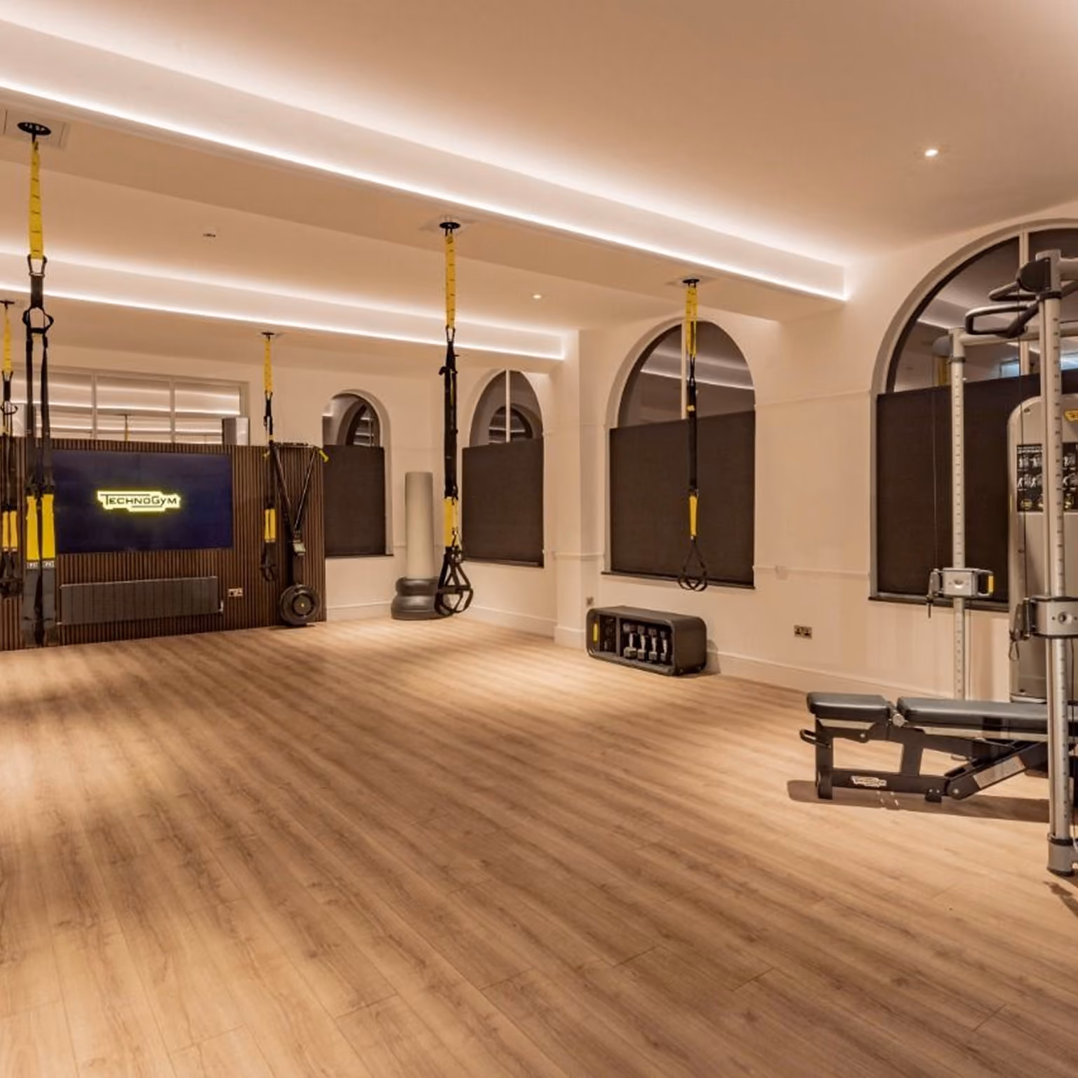 Spacious, modern gym room with wood flooring, black wall panels, and multiple suspension training straps hanging from the ceiling, along with a weight bench and equipment rack under arched windows with black blinds.