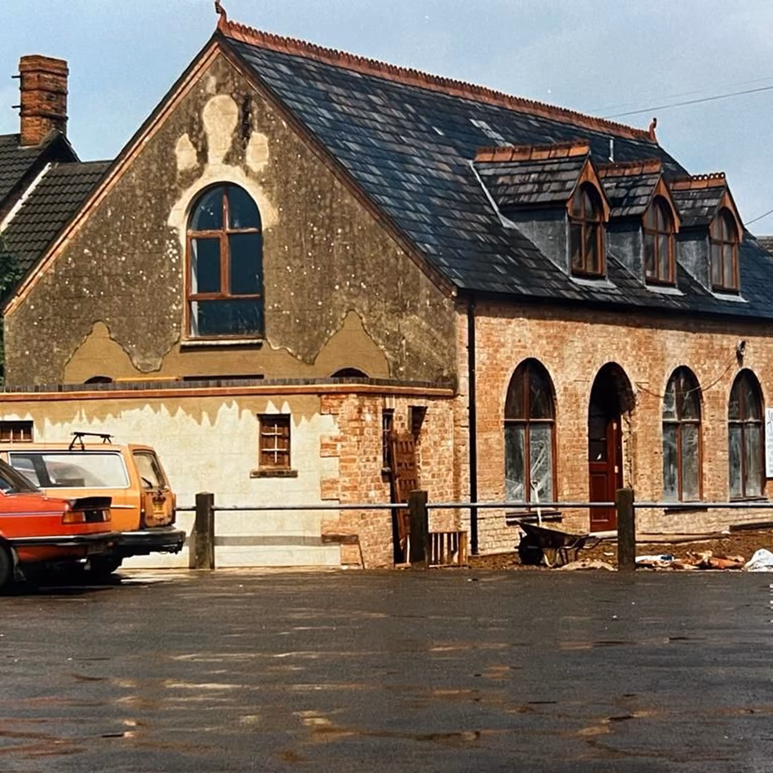 An old building with a slate roof featuring three small dormer windows and arched windows on the brick wall below, a red door, and peeling plaster revealing brick underneath; two vintage cars are parked nearby on a wet asphalt surface.