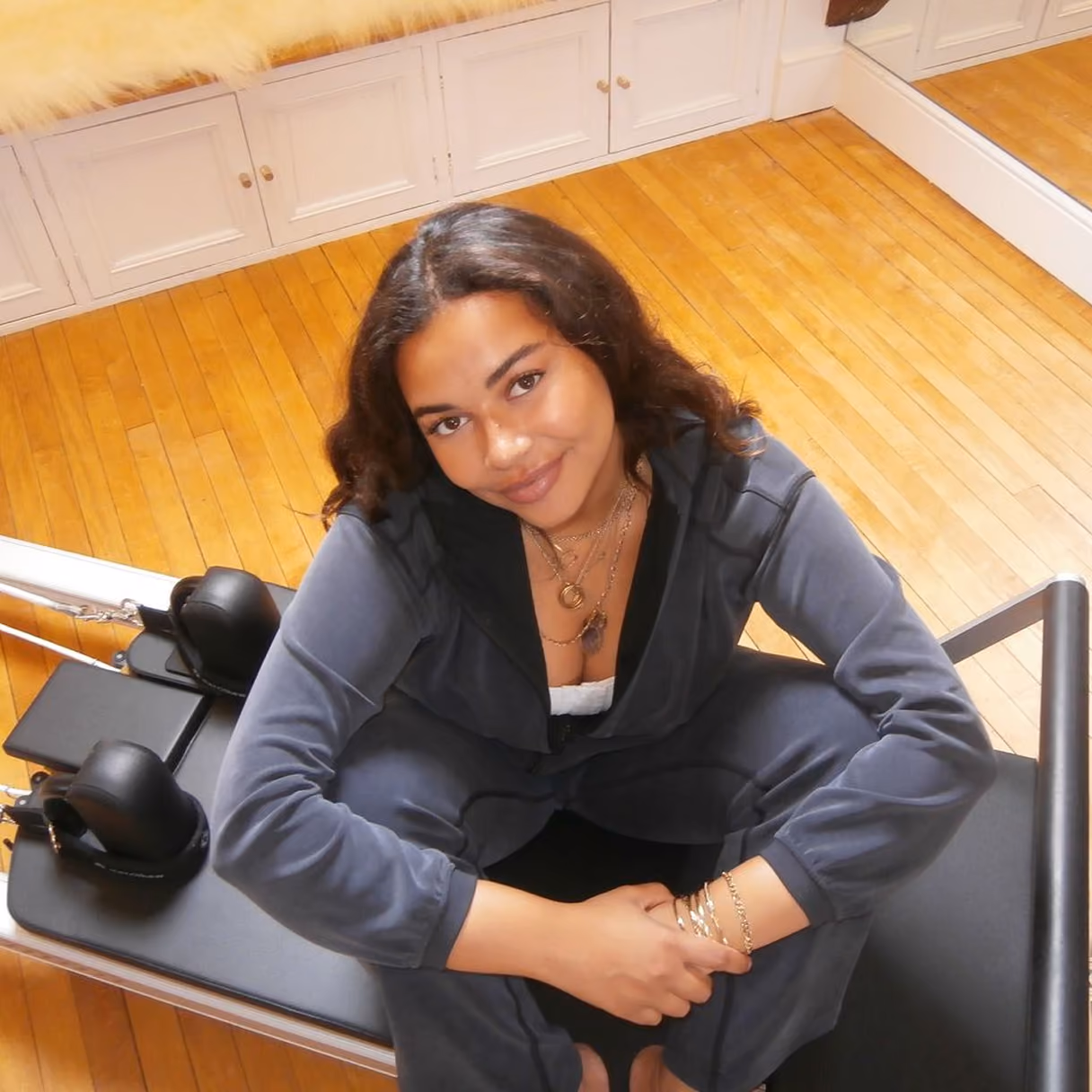 Young woman with long curly dark hair wearing a dark gray tracksuit sitting cross-legged on a Pilates reformer machine in a room with wooden floors and white cabinets.