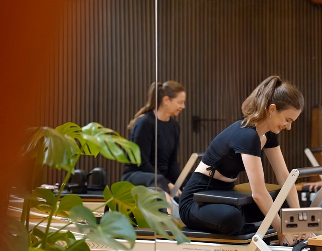 Young woman in black workout clothes using a Pilates reformer machine in a fitness studio with plants and a mirror reflecting her image.
