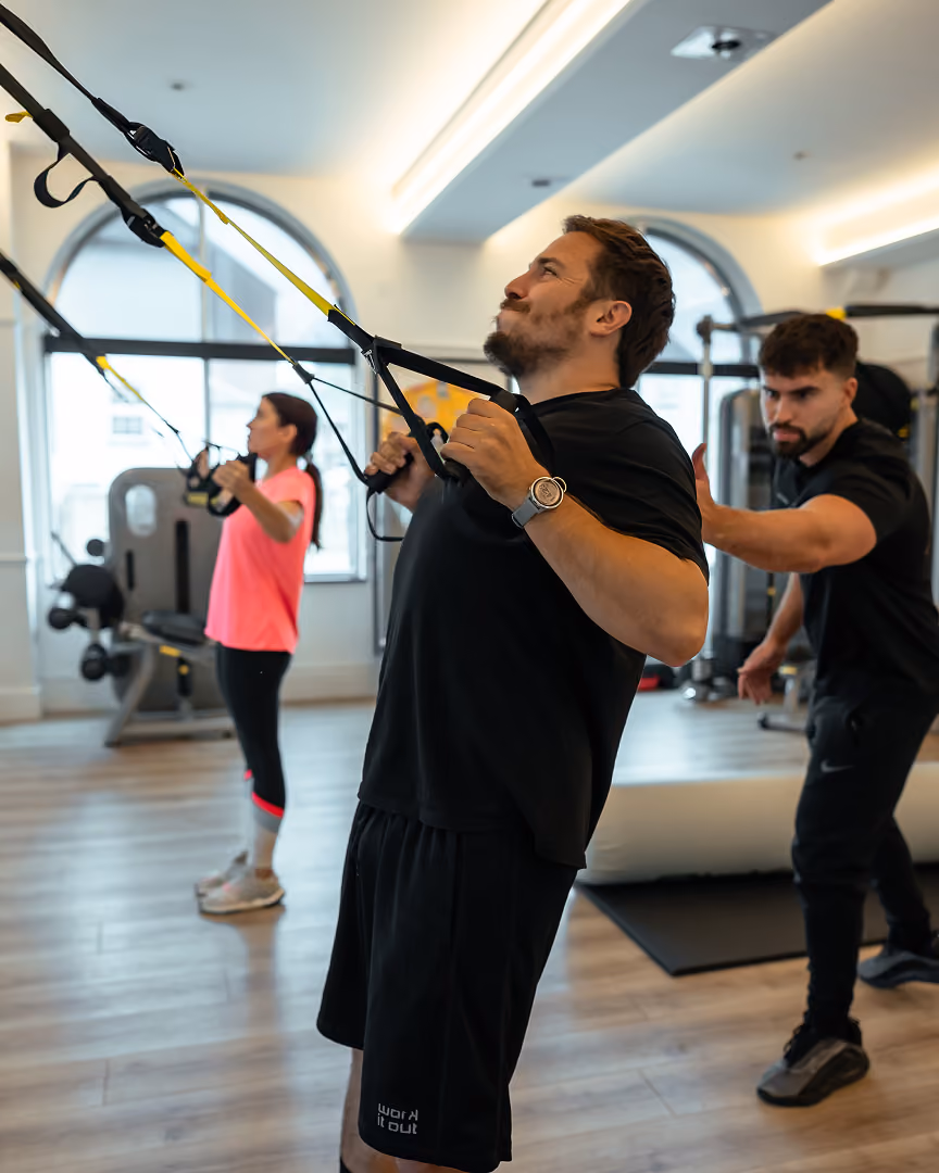 Man in black workout clothes using TRX suspension straps for upper body exercise in a gym while a trainer spots him and a woman in pink workout clothes exercises in the background.