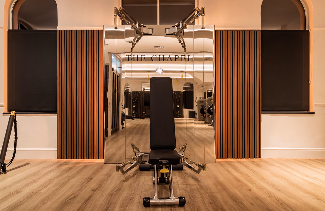 Modern gym machine with black padded seat and backrest in front of mirrored panels labeled 'THE CHAPEL,' with wooden floor and black window coverings in a warmly lit fitness studio.