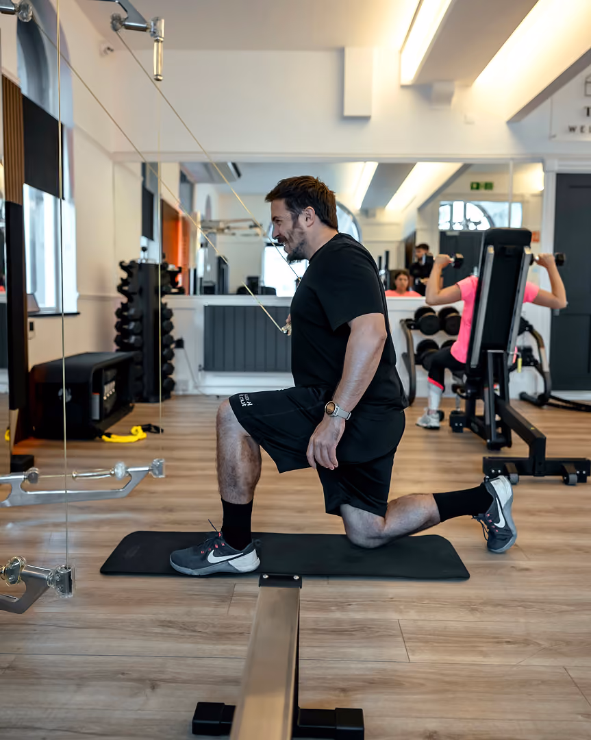 Man in black workout clothes performing a lunge stretch on a black exercise mat in a gym, with another person working out in the background using weight machines.