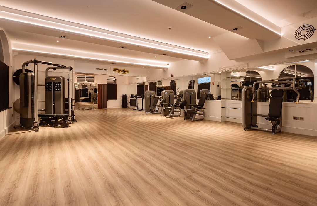 Spacious indoor gym with wooden flooring, ceiling lights, mirrors, and various strength training machines along the walls.
