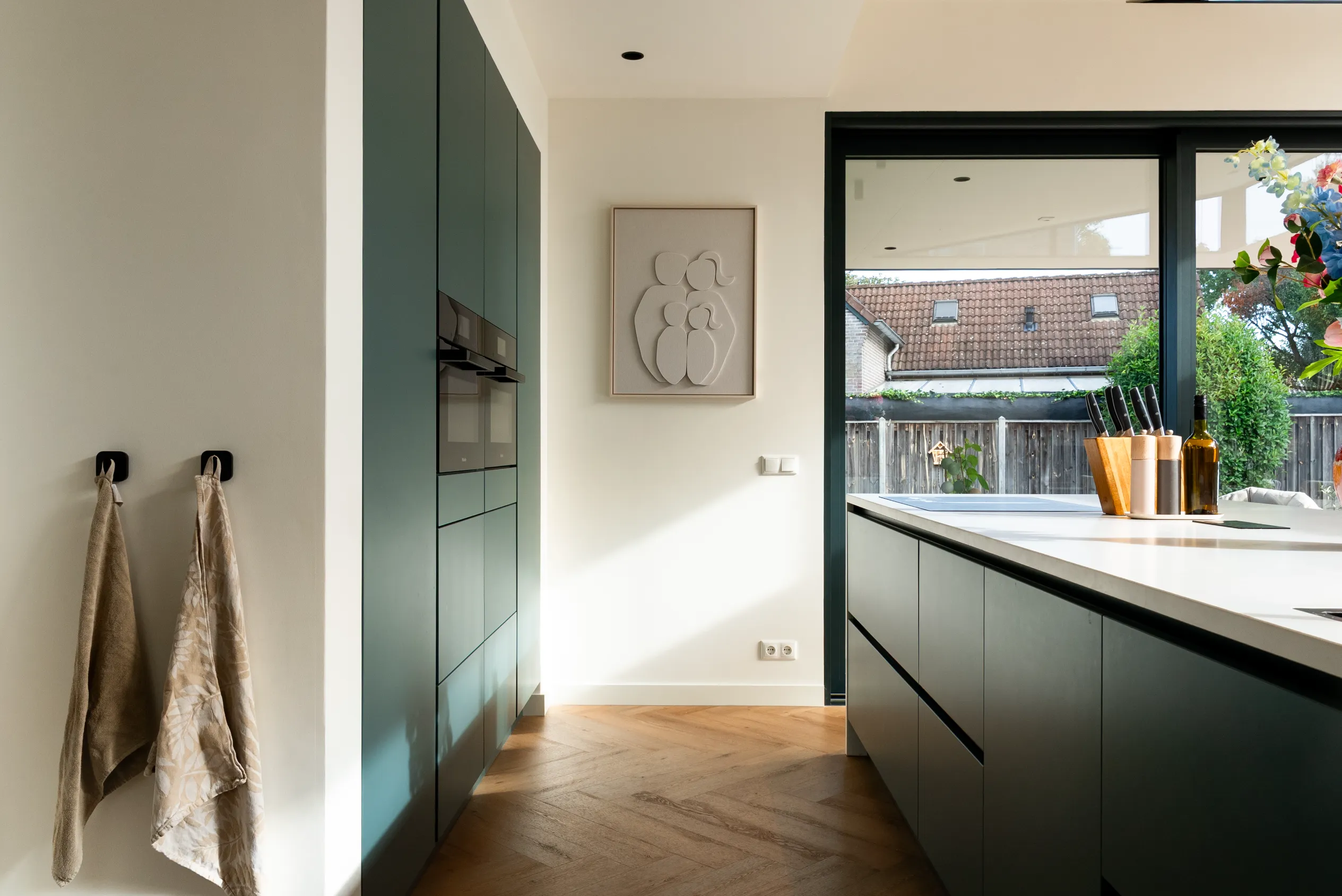 Doorkijk langs de groene keukenkasten naar het aanrechtblad en de tuin