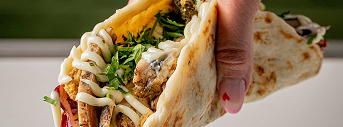 Hand holding pita bread stuffed with fries, greens, and drizzled white sauce.