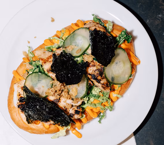 Open sandwich with grilled chicken, cucumber slices, lettuce, seaweed, and orange sauce on white plate.