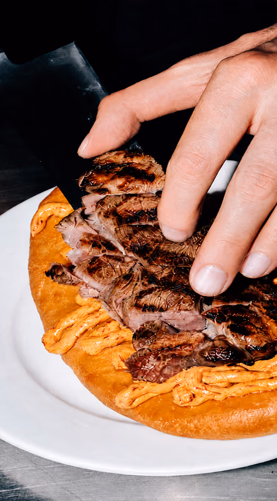 Hand holding sliced grilled steak over a flatbread topped with spicy sauce on a white plate.