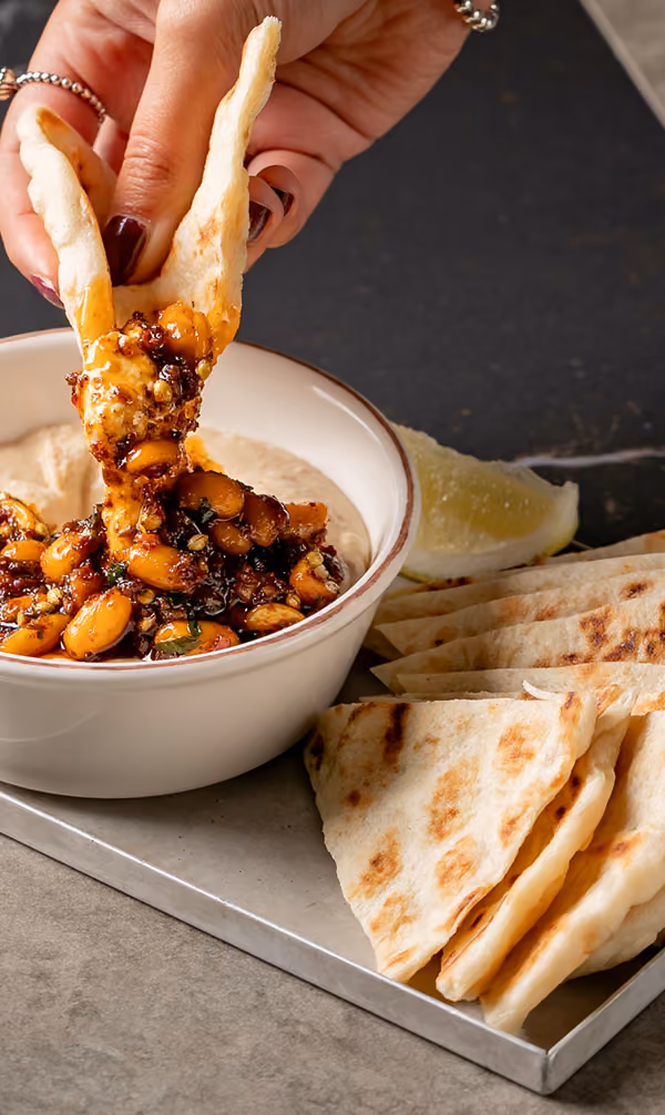 Hand dipping flatbread into a bowl of hummus topped with spicy roasted chickpeas and herbs.