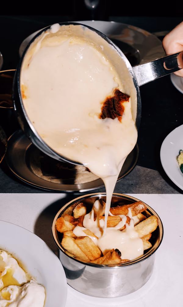 Creamy cheese sauce being poured from a pan onto crispy French fries in a metal cup.