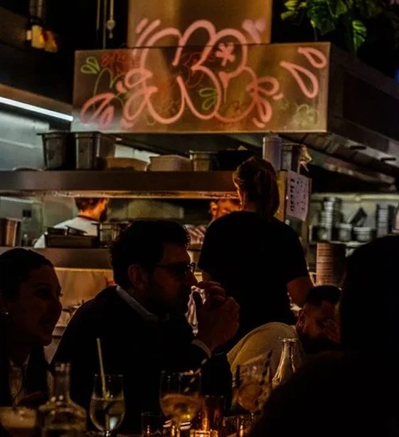 Diners below a pink neon sign by the open kitchen