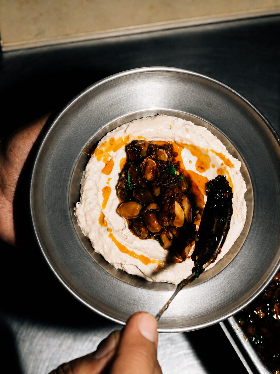A steel bowl of hummus with a spiced topping, held at the pass