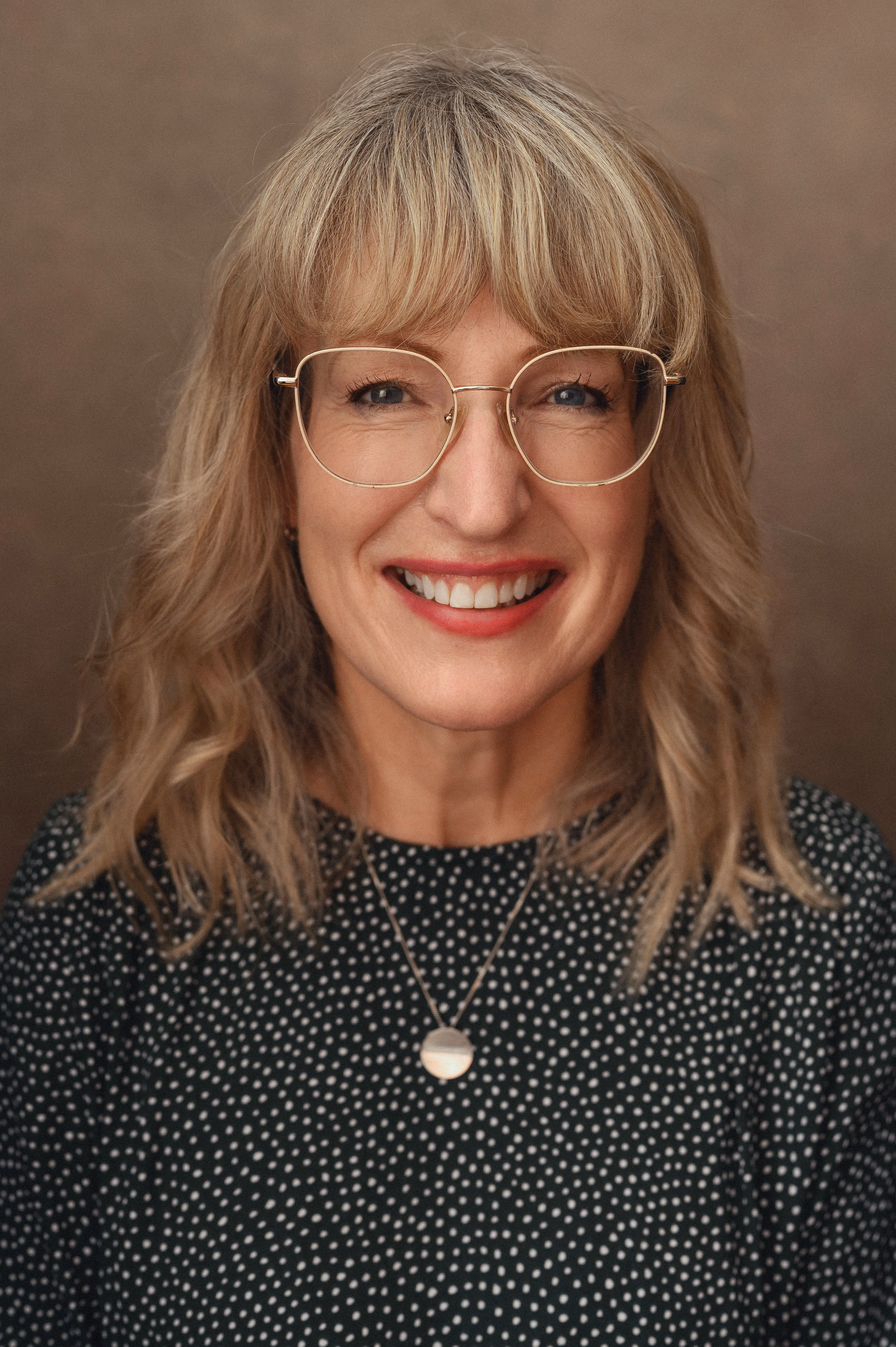 Smiling woman with blonde hair, glasses, and a polka-dot top against a brown background.