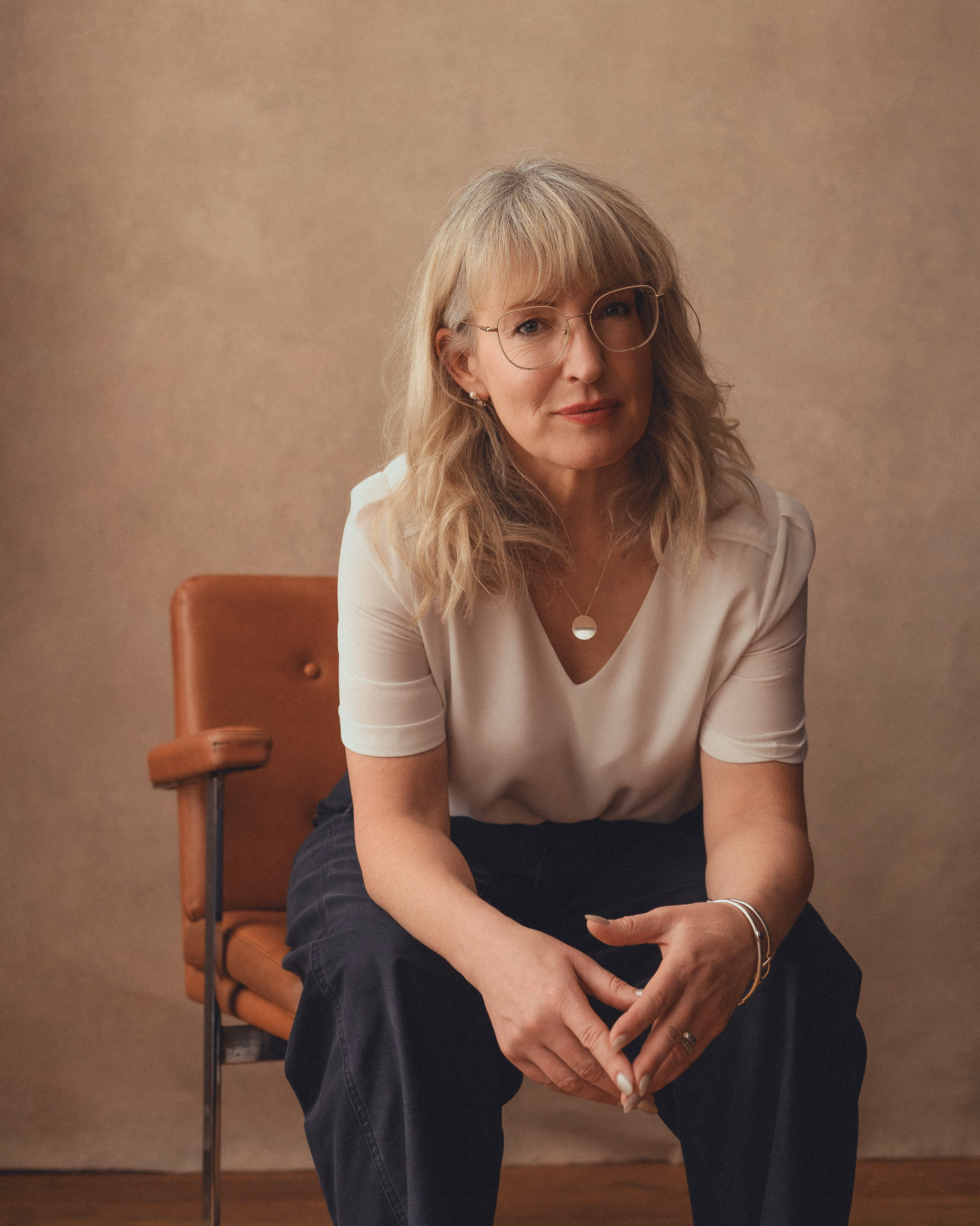 Blonde woman with glasses sitting on a brown chair against a beige backdrop.