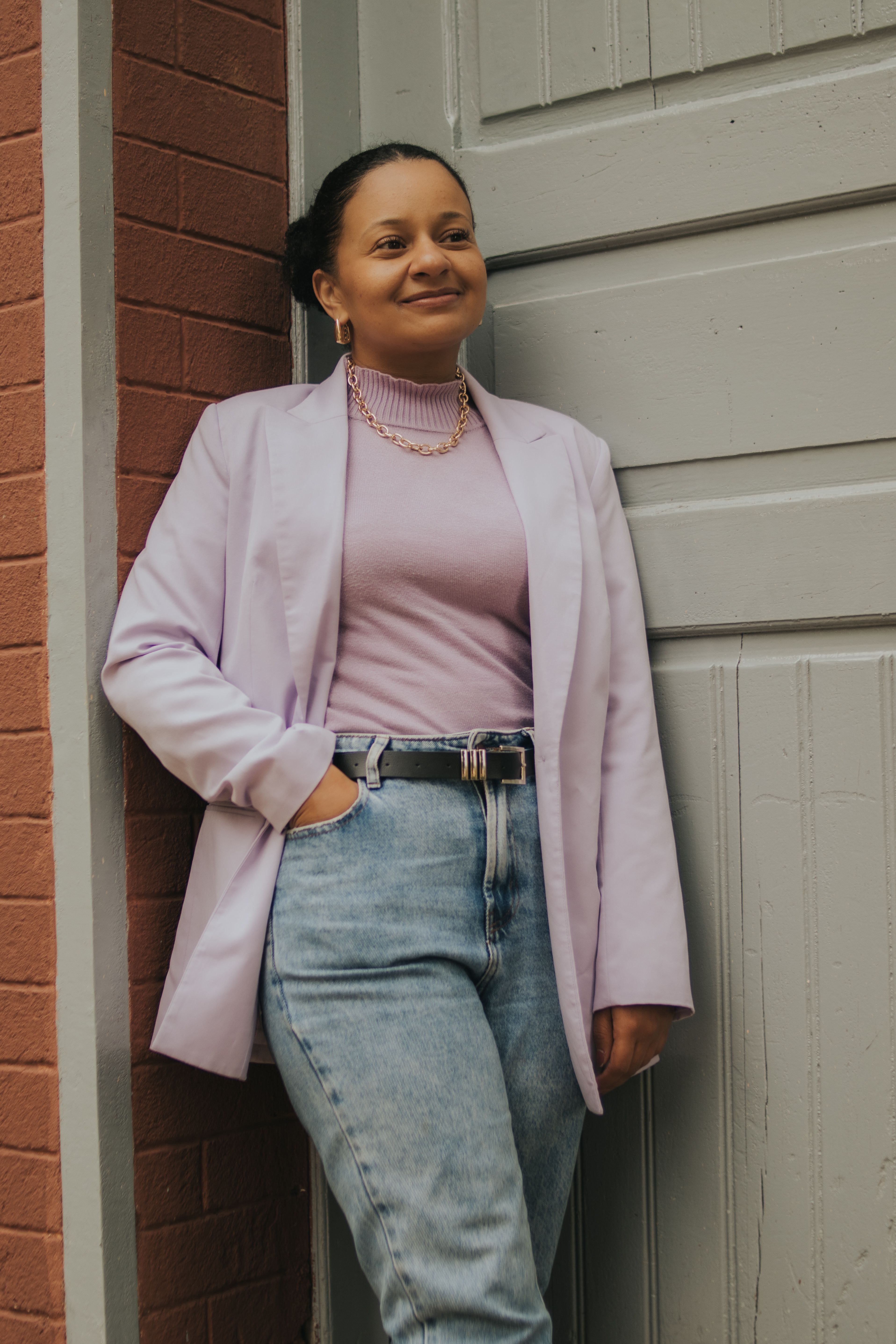 Smiling woman leaning against a brick wall and door, wearing a lavender blazer, top, jeans, and necklace.