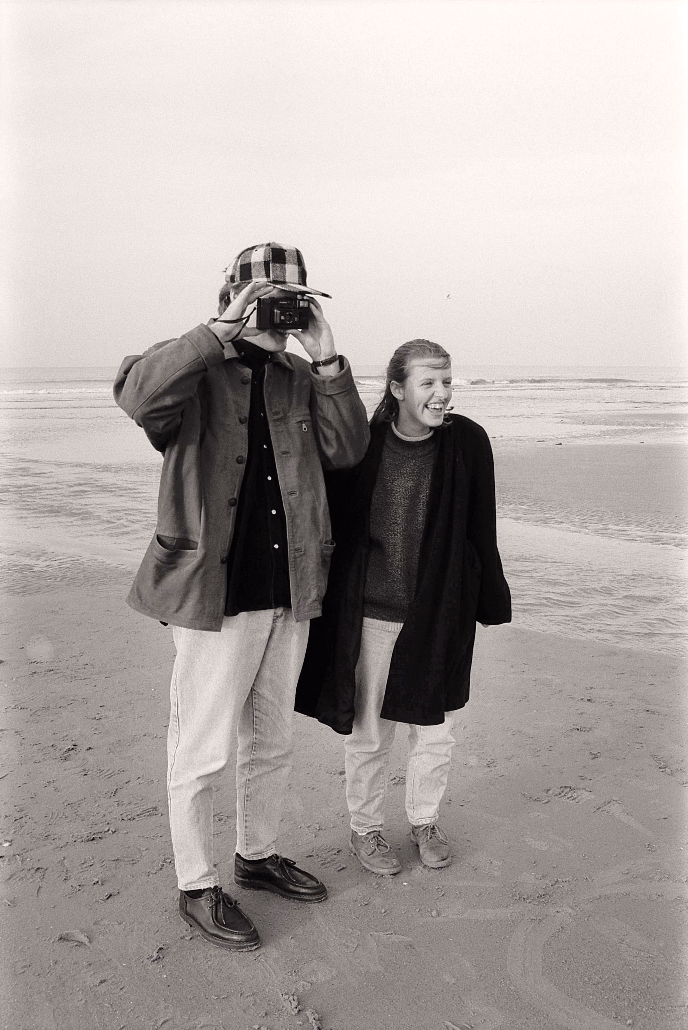 Two people on a beach, one taking a photo with a camera, the other smiling beside them.