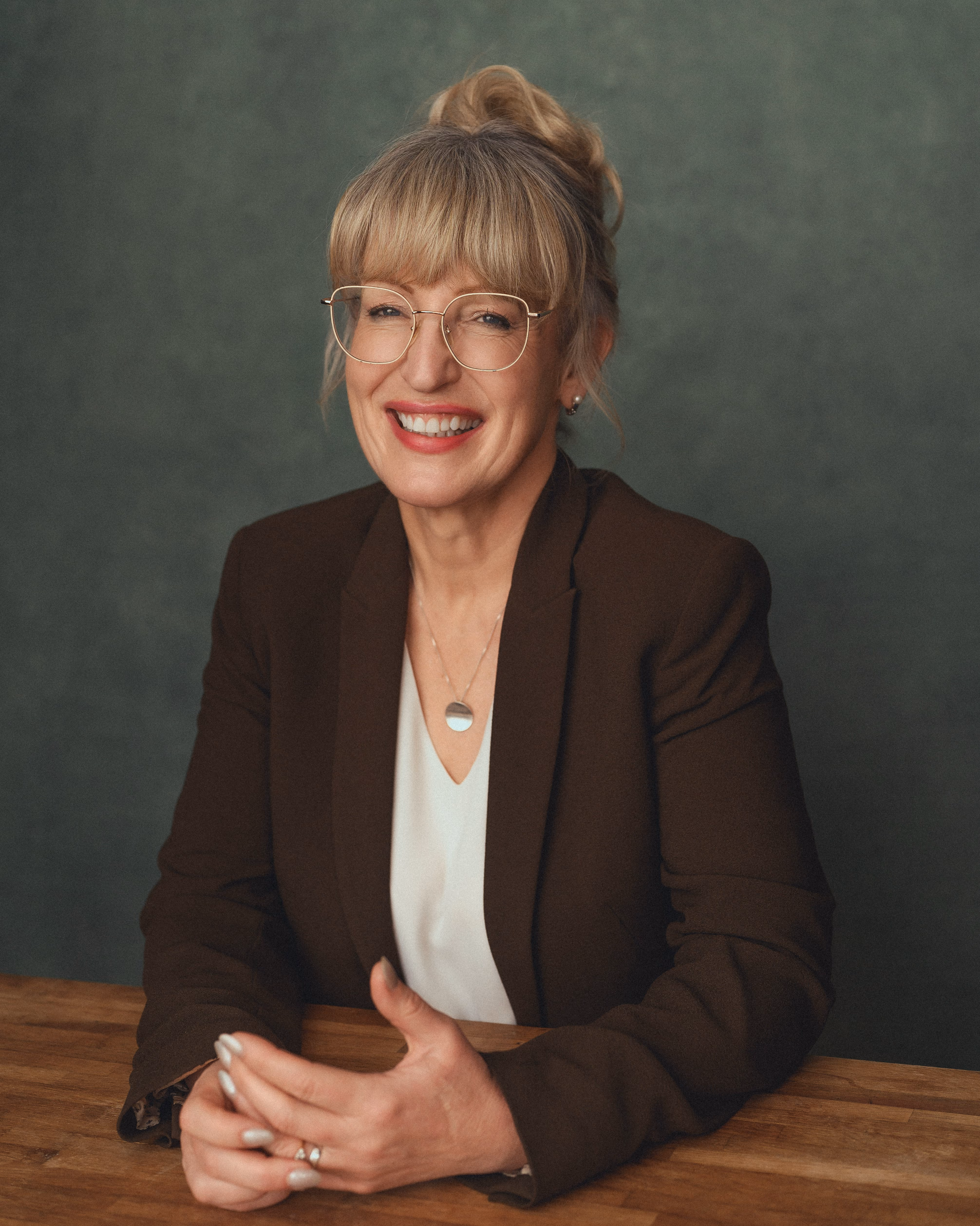 Smiling woman with glasses and blonde hair in a bun, wearing a brown blazer and white blouse.