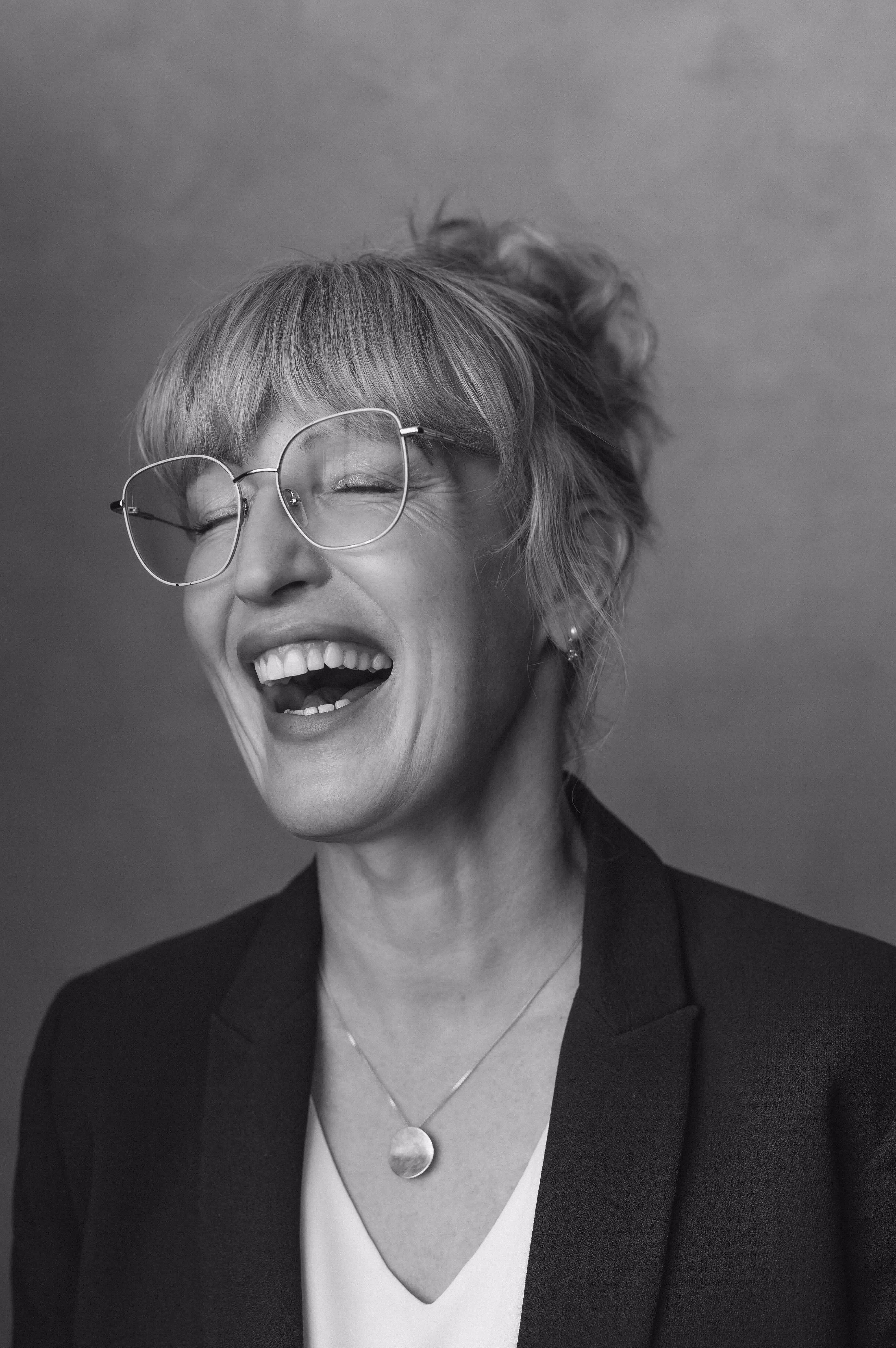 Woman with glasses laughing with eyes closed, wearing a necklace and blazer in black and white portrait.