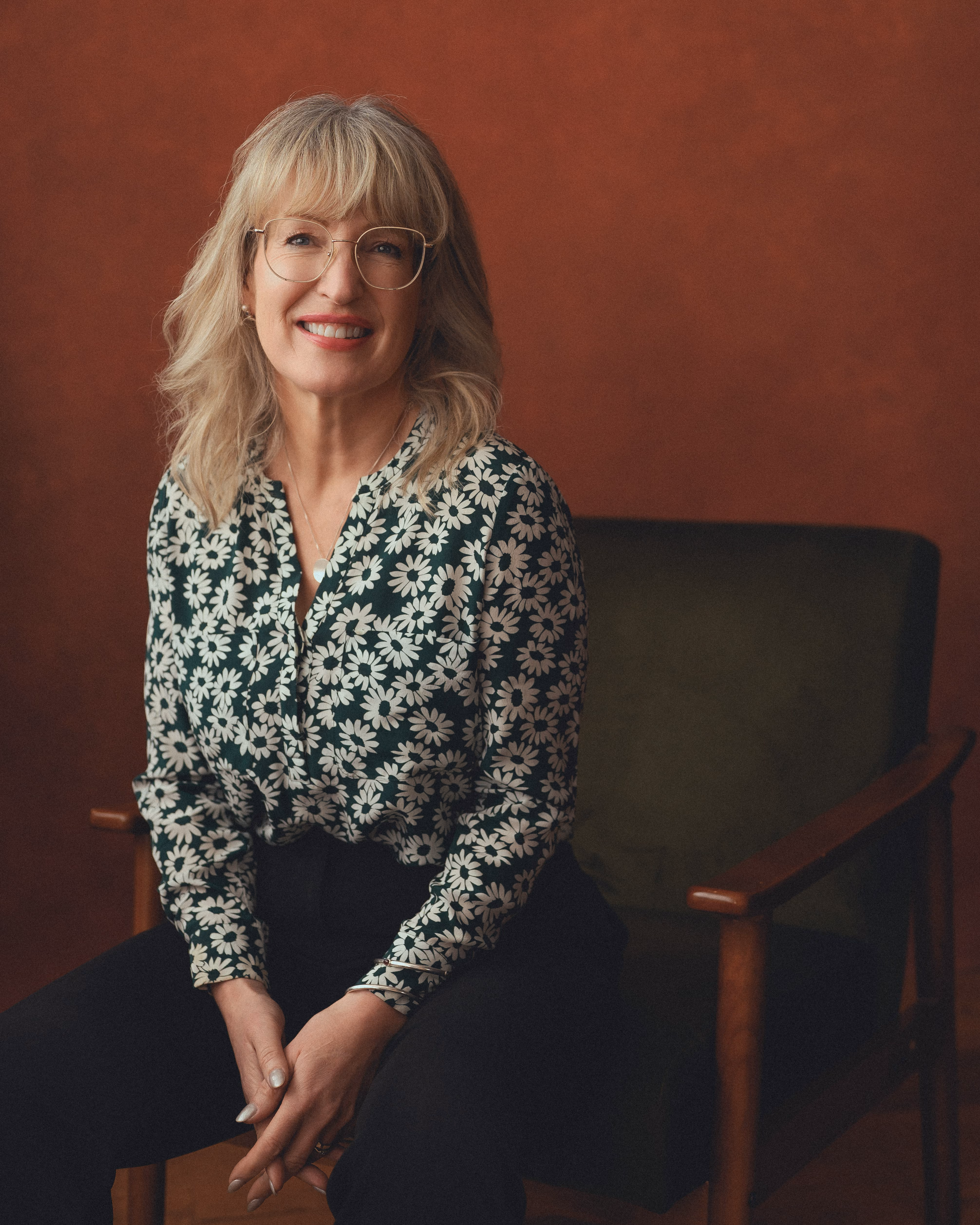 Smiling woman with glasses wearing a green floral blouse seated on a dark chair with a brown background.