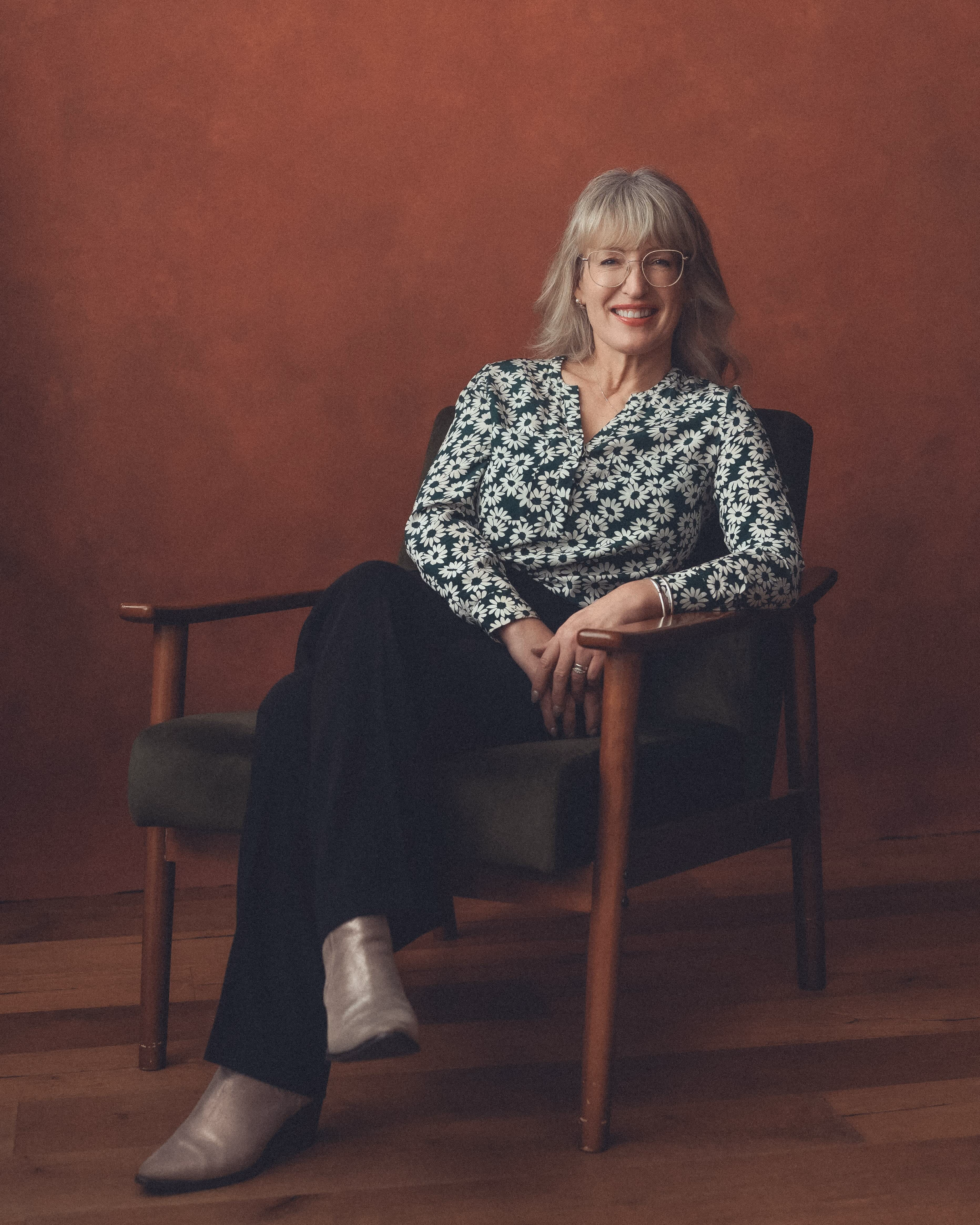 Smiling woman with glasses and floral top sitting cross-legged in a wooden armchair against brown wall.