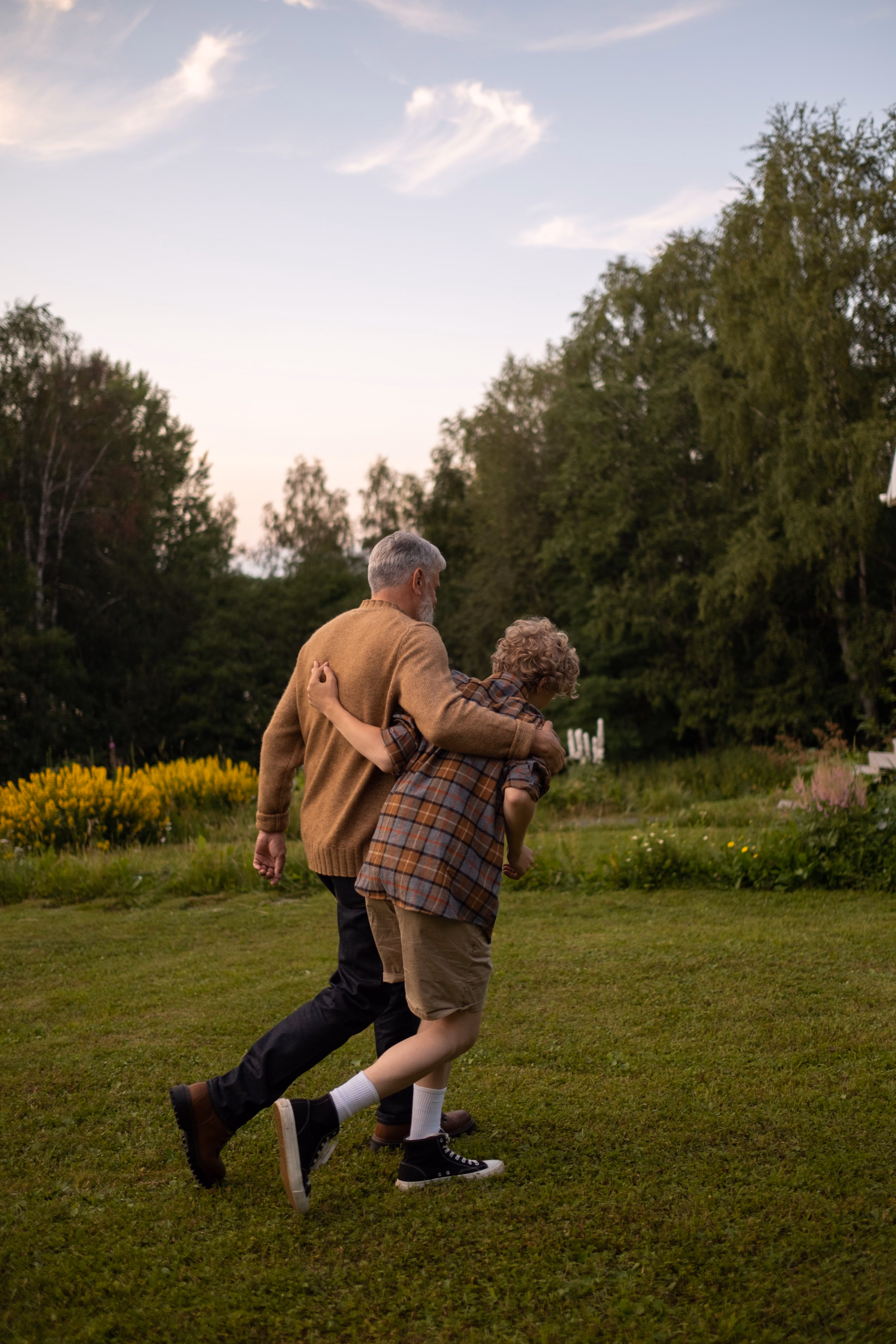 Older man and boy walking arm in arm on grass with trees and flowers in the background.