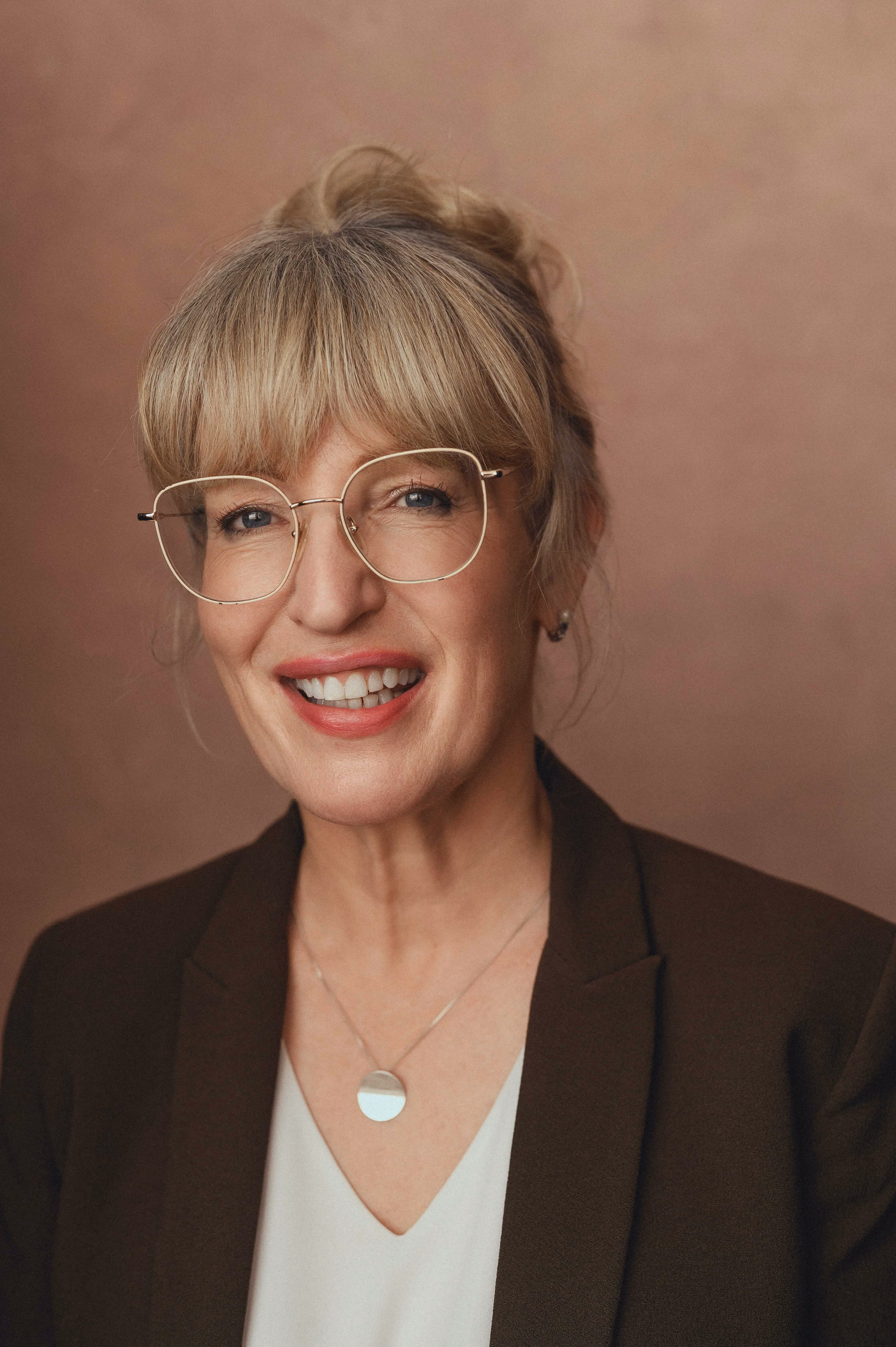 Smiling older woman with blonde hair, glasses, and a dark blazer against a beige background. A psychosexual therapist based in London offering sessions online.